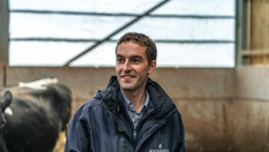 Grant connor looks into middle distance in cattle shed - cow in background over his right shoulder